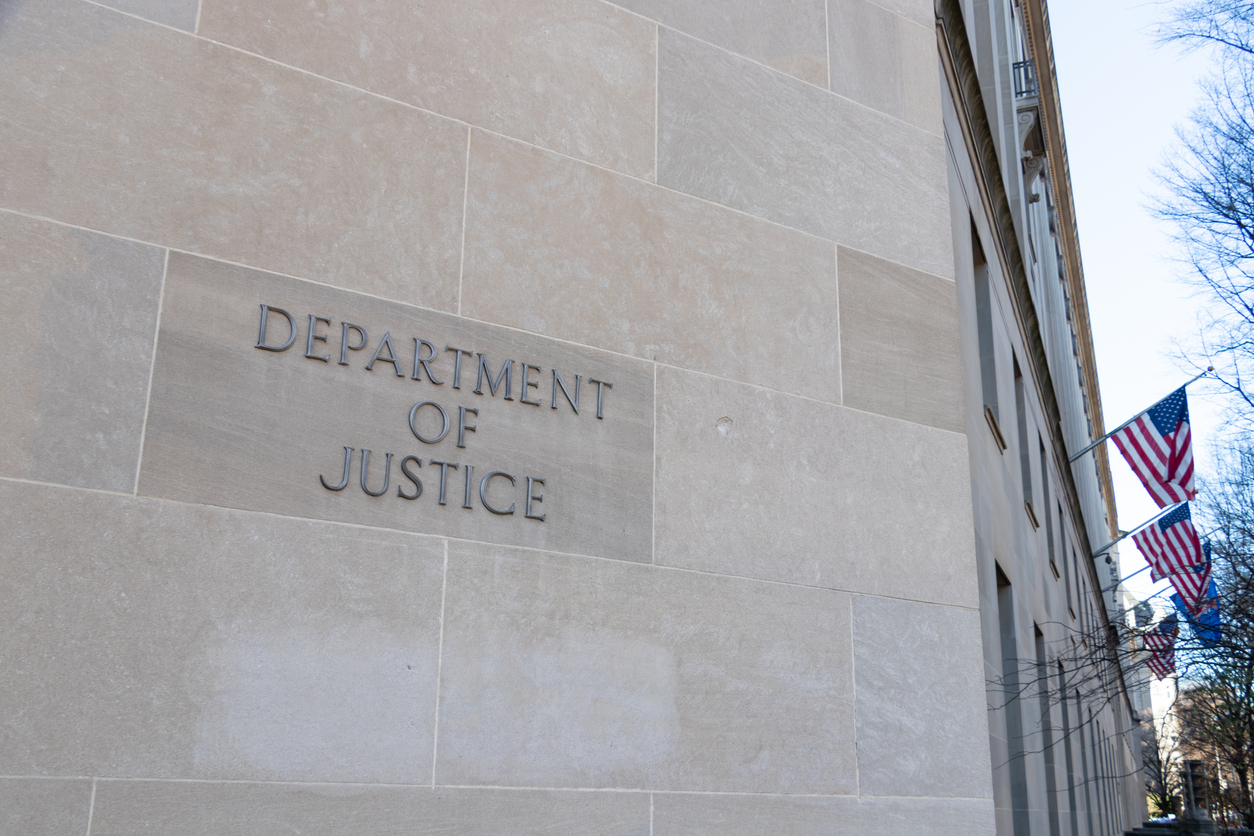 Department of Justice text on the side of their HQ in Washington, D.C. - American flags in the background.